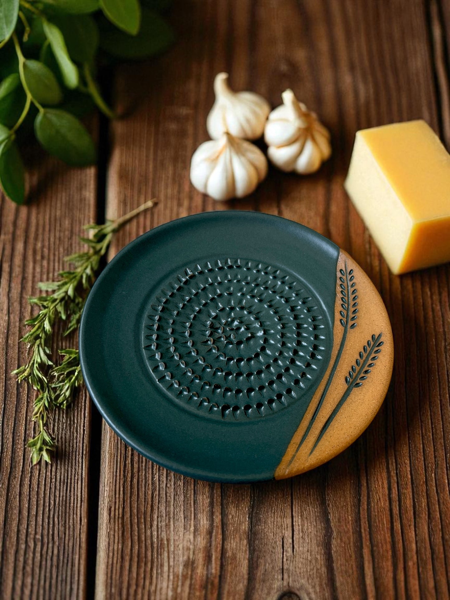 ceramic grater on a wooden table with a block of cheese, garlic cloves, and fresh herbs