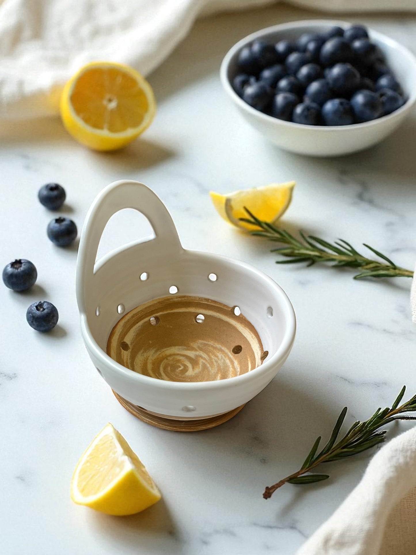 White ceramic colander with wooden base on a marble surface with lemons and blueberries.