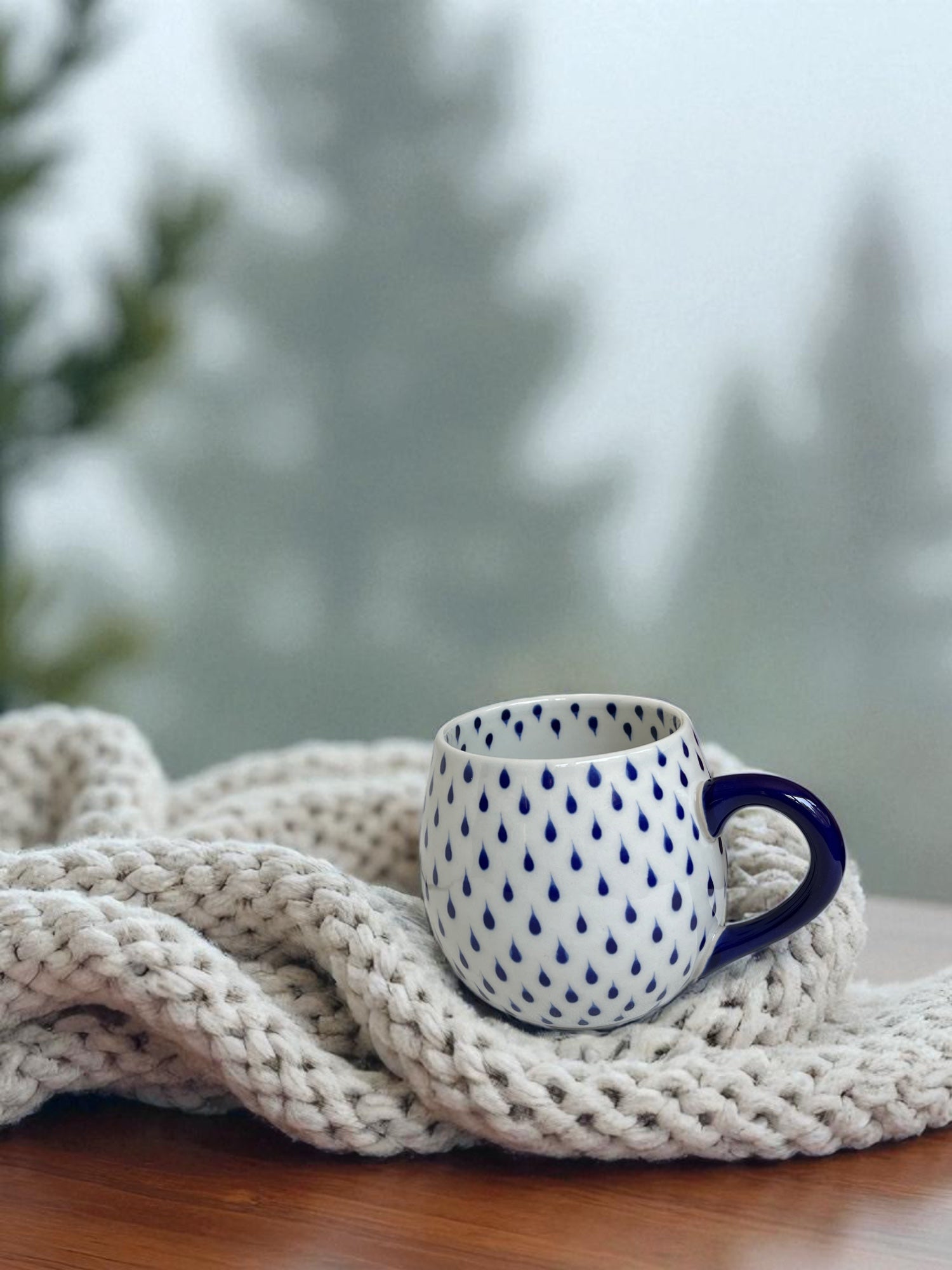 white glazed mug with blue raindrops rests on knitted blanket with evergreen trees visible out the window