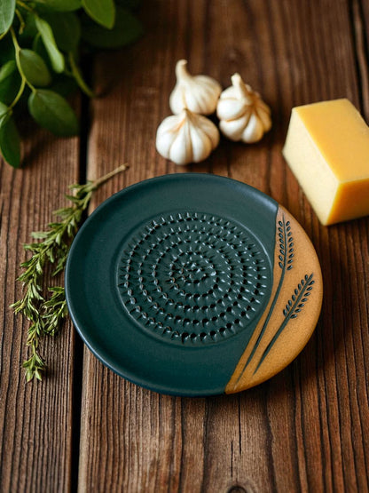 ceramic grater on a wooden table with a block of cheese, garlic cloves, and fresh herbs