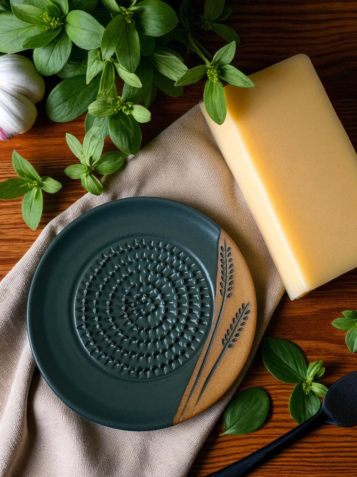 ceramic grater on a linen cloth on a wooden table with a block of cheese, garlic cloves, and fresh herbs