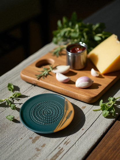 ceramic grater on a wooden table with a cutting board with ablock of cheese, garlic cloves, and fresh herbs