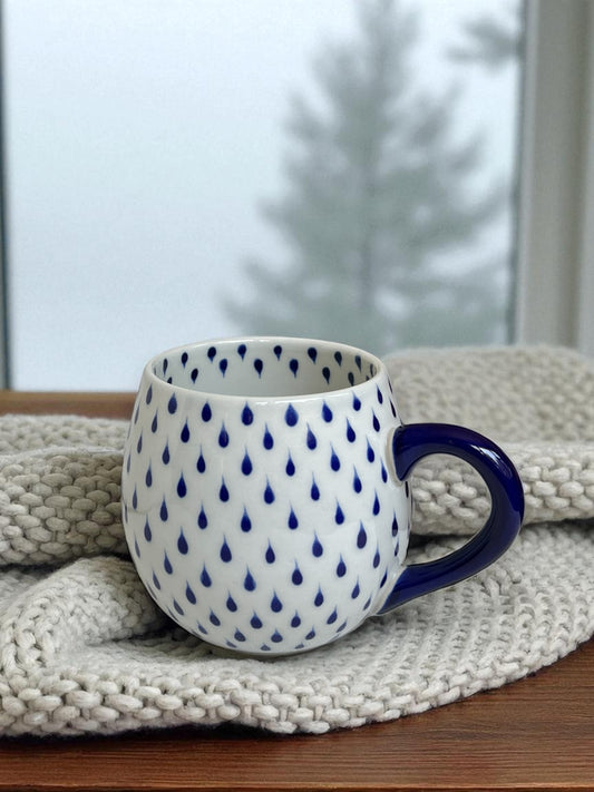 white glazed mug with blue raindrops rests on knitted blanket with evergreen trees visible out the window